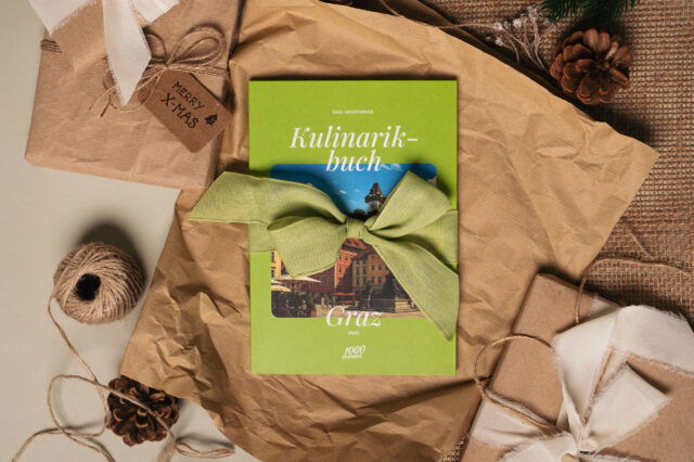 Das 1000things Kulinarikbuch Graz liegt weihnachtlich dekoriert auf Packpapier und ist mit einer grünen Schleife verpackt.