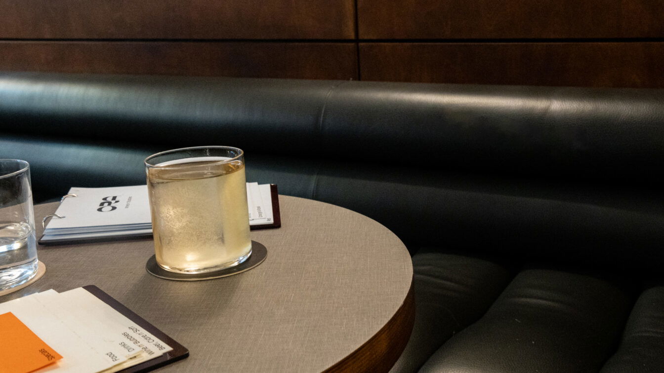(c) Sonja Koller | 1000things Detailaufnahme eines stilvollen Bartischs der Ora Bar in Zürich: ein Glas mit hellem Drink auf einem runden Tisch, daneben Wasserglas und Getränkekarten auf einer eleganten Lederbank – minimalistische, moderne Atmosphäre typisch für Zürcher Bars.