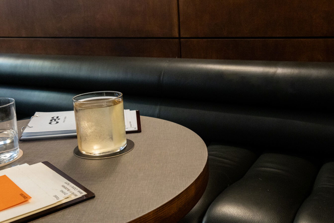 Detailaufnahme eines stilvollen Bartischs der Ora Bar in Zürich: ein Glas mit hellem Drink auf einem runden Tisch, daneben Wasserglas und Getränkekarten auf einer eleganten Lederbank – minimalistische, moderne Atmosphäre typisch für Zürcher Bars.