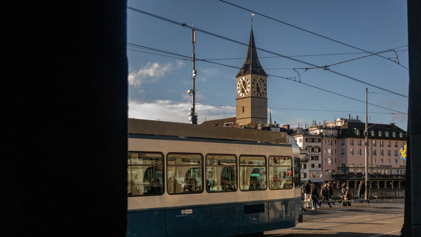 (c) Sonja Koller | 1000things Straßenszene in Zürich mit einer vorbeifahrenden blauen Tram im Vordergrund und dem Turm der St. Peterskirche mit ihrer großen Uhr im Hintergrund, aufgenommen bei klarem Wetter am späten Nachmittag.