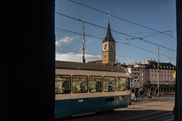 (c) Sonja Koller | 1000things Straßenszene in Zürich mit einer vorbeifahrenden blauen Tram im Vordergrund und dem Turm der St. Peterskirche mit ihrer großen Uhr im Hintergrund, aufgenommen bei klarem Wetter am späten Nachmittag.