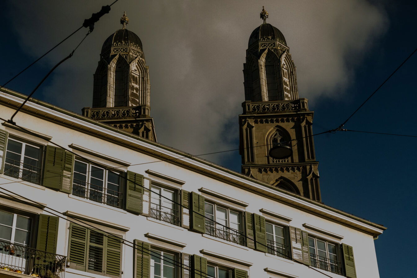 Gratis in Zürich: Blick auf die beiden Türme des Grossmünsters über den grünen Fensterläden eines Wohnhauses vor dramatischem Himmel.