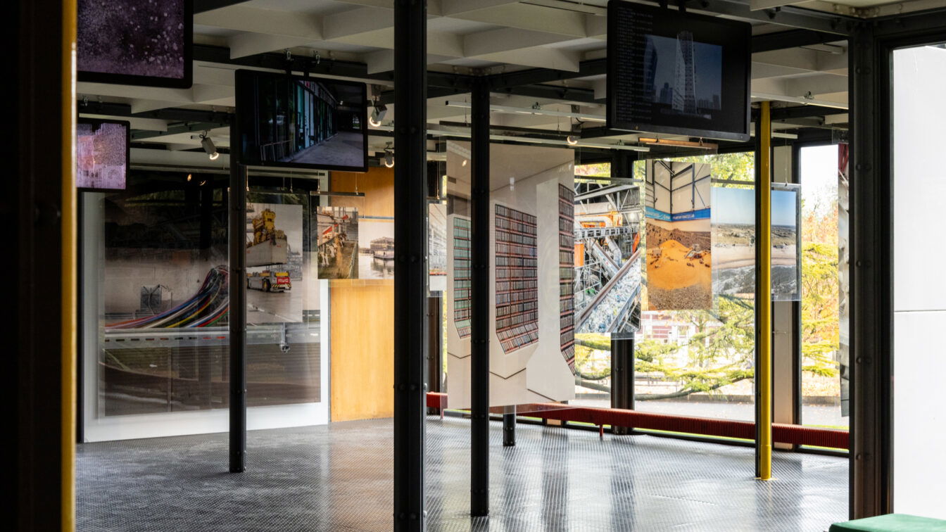 Blick in einen modernen Ausstellungsraum mit großen Fenstern, durch die herbstliche Bäume zu sehen sind. Zwischen schwarzen Metallstützen hängen Fotografien und Bildschirme mit urbanen und industriellen Motiven. Der Raum hat einen dunklen Metallboden und klare, minimalistische Architektur mit Stahl- und Glaselementen.
