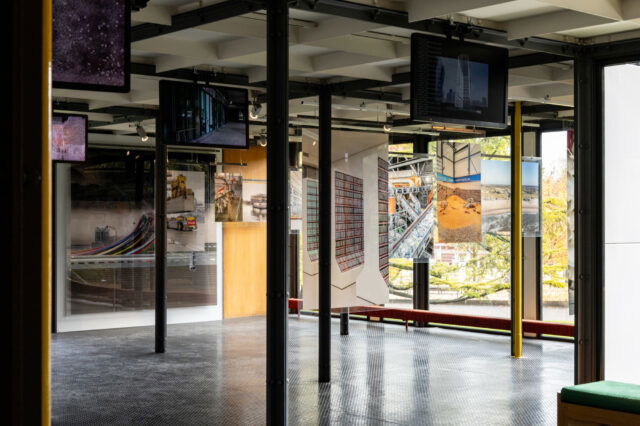 Blick in einen modernen Ausstellungsraum mit großen Fenstern, durch die herbstliche Bäume zu sehen sind. Zwischen schwarzen Metallstützen hängen Fotografien und Bildschirme mit urbanen und industriellen Motiven. Der Raum hat einen dunklen Metallboden und klare, minimalistische Architektur mit Stahl- und Glaselementen.