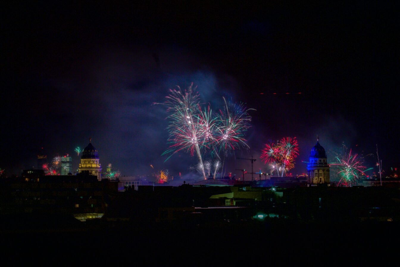 Nächtliche Skyline von Berlin an Silvester: Vor dunklem Himmel erhellen bunte Feuerwerksfontänen und Funkenexplosionen die Stadt. Links und rechts ragen zwei angestrahlte Kuppelbauten hervor, während im Hintergrund weitere Gebäude und Kräne schemenhaft sichtbar sind. Rauch und farbige Lichtspuren breiten sich über der Stadt aus.