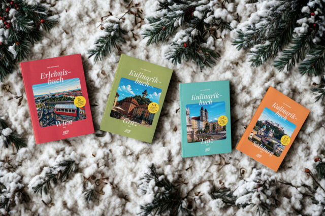 Vier 1000things Bücher – das Erlebnisbuch Wien sowie die Kulinarikbücher Graz, Linz und Salzburg – liegen auf schneebedeckten Tannenzweigen im winterlichen Setting.