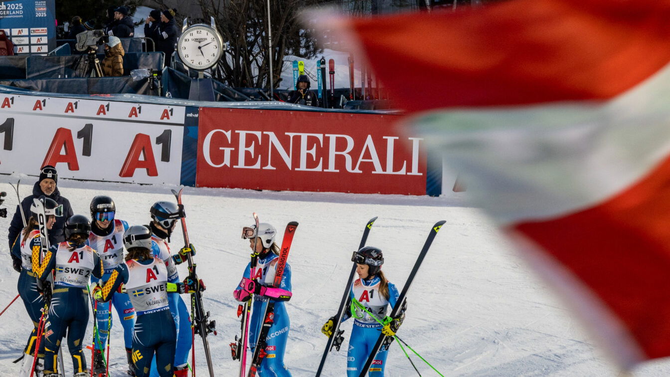 „Gruppe von Skirennläuferinnen in Rennanzügen und Helmen steht nach einem Wettkampf auf der Skipiste zusammen. Im Hintergrund sind Werbebanner, ein großer Standuhr und Zuschauer zu sehen; im Vordergrund weht unscharf eine rot-weiße Fahne ins Bild.