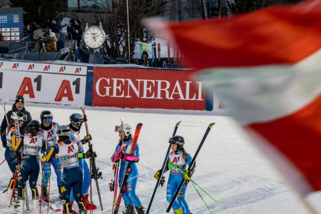 „Gruppe von Skirennläuferinnen in Rennanzügen und Helmen steht nach einem Wettkampf auf der Skipiste zusammen. Im Hintergrund sind Werbebanner, ein großer Standuhr und Zuschauer zu sehen; im Vordergrund weht unscharf eine rot-weiße Fahne ins Bild.