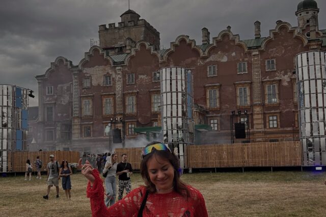 Person in rotem, grob gehäkeltem Oberteil tanzt lachend auf einem Open-Air-Techno-Festival vor einer verwitterten historischen Backsteinfassade; umgeben von Festivalbesucherinnen, Bühnenaufbauten und grauem, dramatischem Himmel.