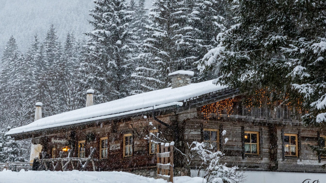 Ein verschneites Berghütten-Panorama in Österreich im Winter: Eine rustikale Holzhütte mit warm leuchtenden Fenstern und Lichterketten steht am Waldrand, umgeben von schneebedeckten Tannen und dicht fallendem Schnee.