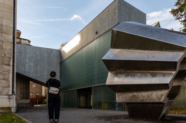 Eine Person steht vor einem modernen Museumsgebäude mit klaren geometrischen Formen, grünlich getönten Glaswänden und einer markanten Metallskulptur im Vordergrund. Das Wechselspiel aus Stein, Glas und Metall verleiht dem Ort eine architektonisch spannende Atmosphäre. Das Bild zeigt Winterthur, der sich gut als Ausflugsziel ab Zürich eignet.