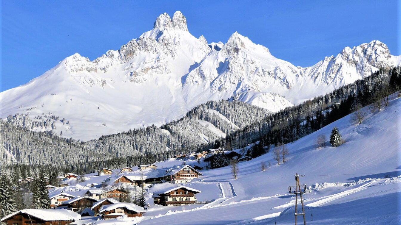 Auf dem Bild sieht man das verschneite Bergdorf Filzmoos mit zahlreichen Holzhäusern, eingebettet in eine Winterlandschaft, überragt vom markanten, schneebedeckten Gipfel der Bischofsmütze.