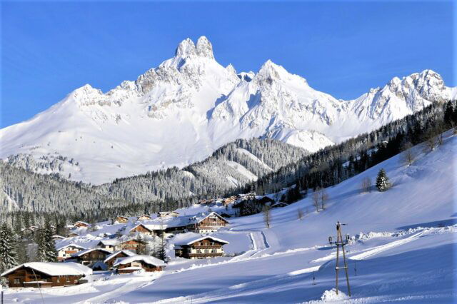 Auf dem Bild sieht man das verschneite Bergdorf Filzmoos mit zahlreichen Holzhäusern, eingebettet in eine Winterlandschaft, überragt vom markanten, schneebedeckten Gipfel der Bischofsmütze.