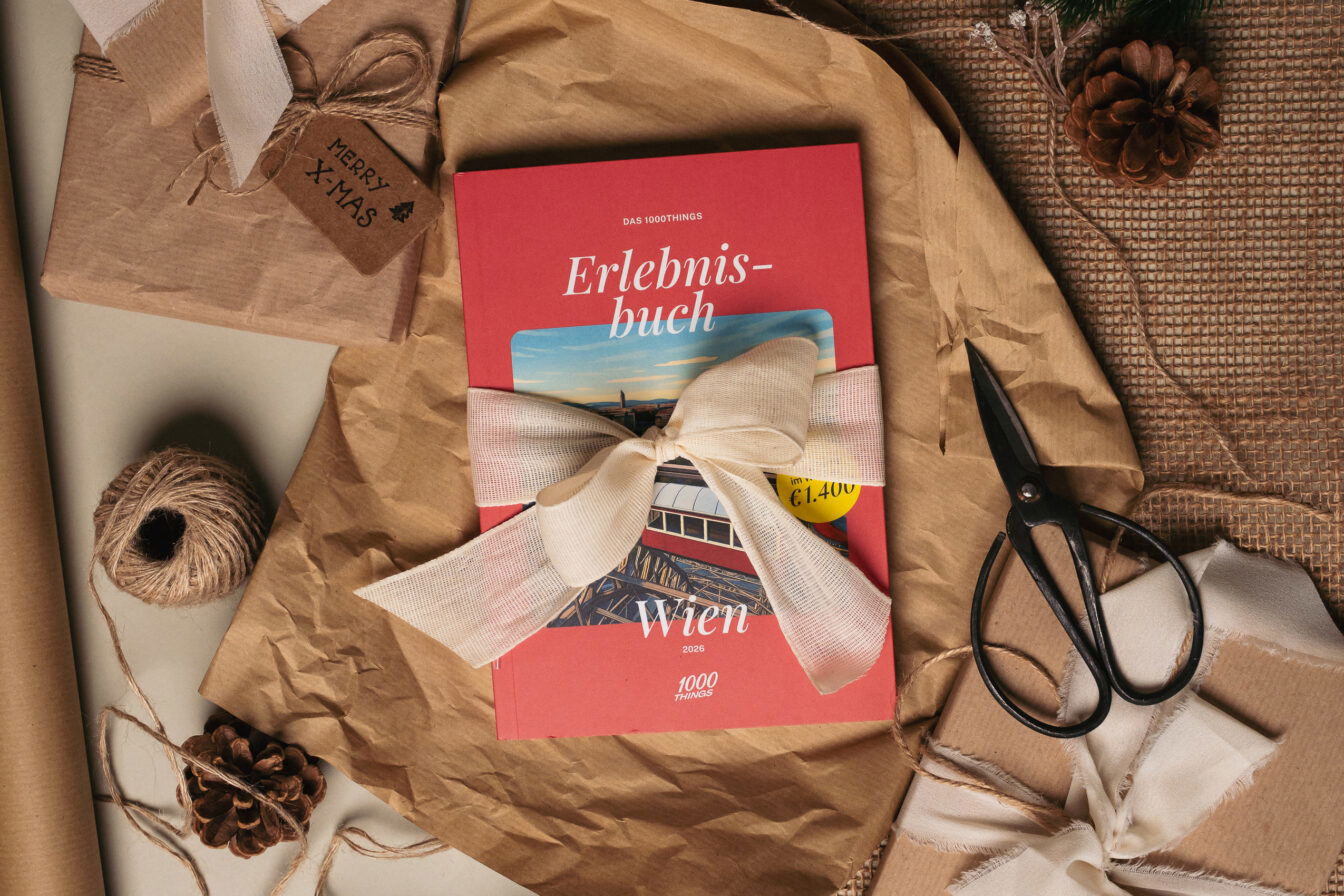 Weihnachtlich dekorierte Szene mit dem roten 1000things Erlebnisbuch Wien 2026, das mit einer großen cremefarbenen Schleife verpackt ist; darum Geschenkpapier, Schnur, Kiefernzapfen und eine Schere.