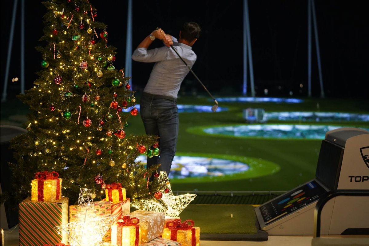 Ein Mann versucht einen Abschlag am Golfplatz. Es ist Abend, im Vordergrund steht ein erleuchteter Weihnachtsbaum mit Geschenken.