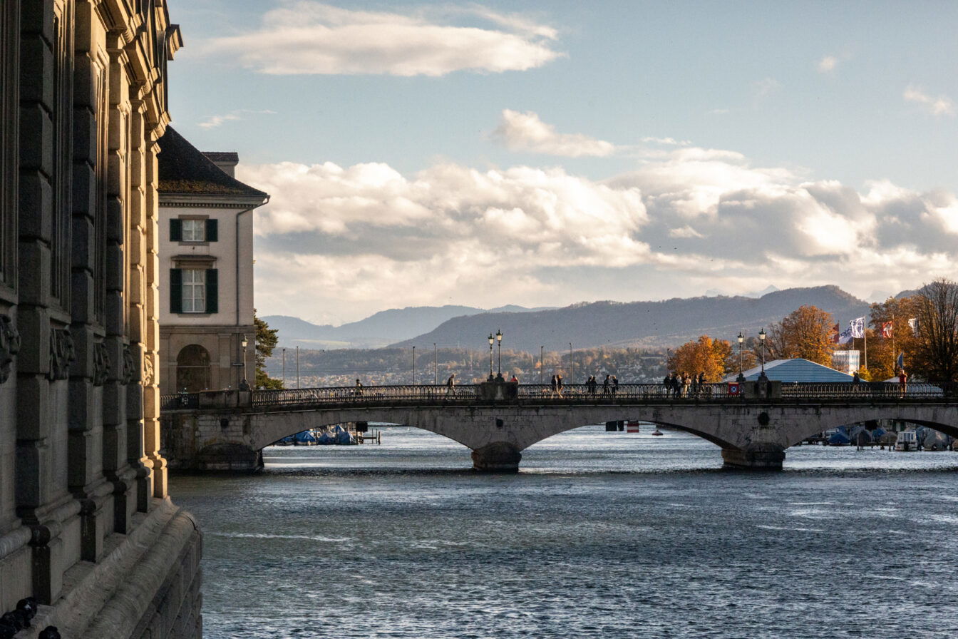 Blick über einen Fluss in Zürich auf eine steinerne Brücke mit Fußgängern, dahinter herbstliche Bäume, Stadtgebäude und Hügel unter einem teils bewölkten Himmel; links im Vordergrund die Fassade eines historischen Gebäudes