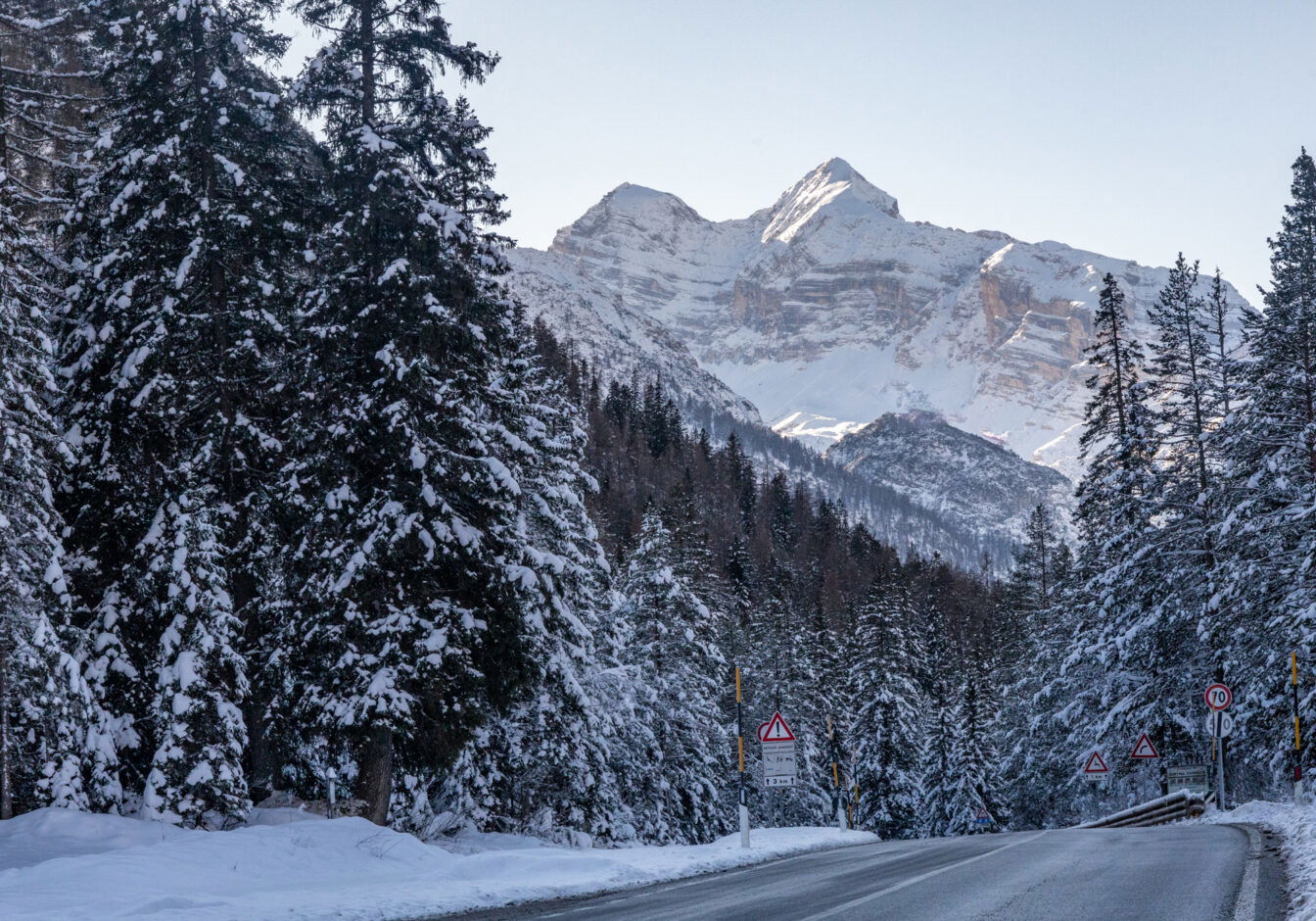 Winterliche Berglandschaft nahe Cortina d’Ampezzo mit schneebedeckten Tannen entlang einer Straße; im Hintergrund erheben sich imposante, sonnige Dolomitengipfel, während Verkehrsschilder die kurvige Strecke markieren.