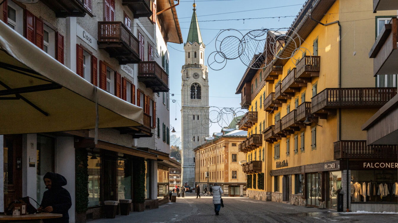 Fußgängerzone in Cortina d’Ampezzo mit traditionellen alpinen Häusern, Holzläden und Balkonen; im Hintergrund ragt der markante Glockenturm der Basilika auf, während einige Menschen die gepflasterte Straße entlanggehen.