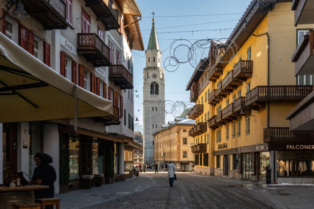 Fußgängerzone in Cortina d’Ampezzo mit traditionellen alpinen Häusern, Holzläden und Balkonen; im Hintergrund ragt der markante Glockenturm der Basilika auf, während einige Menschen die gepflasterte Straße entlanggehen.