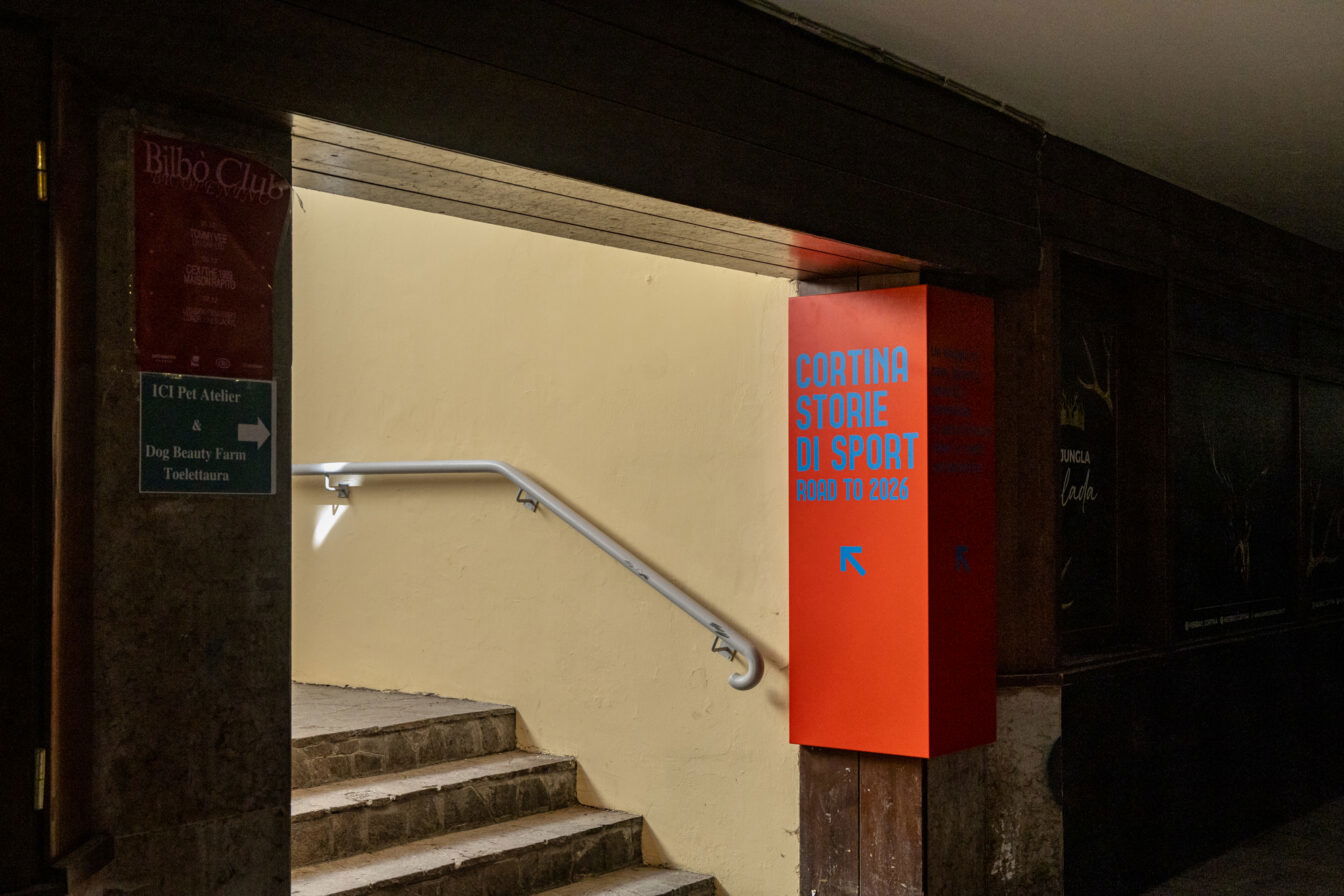 Eingang zu einem unterirdischen Durchgang in Cortina d’Ampezzo; an der Wand steht ein rotes Informationssäulenschild mit der Aufschrift ‚Cortina Storie di Sport – Road to 2026‘, daneben eine Treppe mit Handlauf, die ins Licht führt.