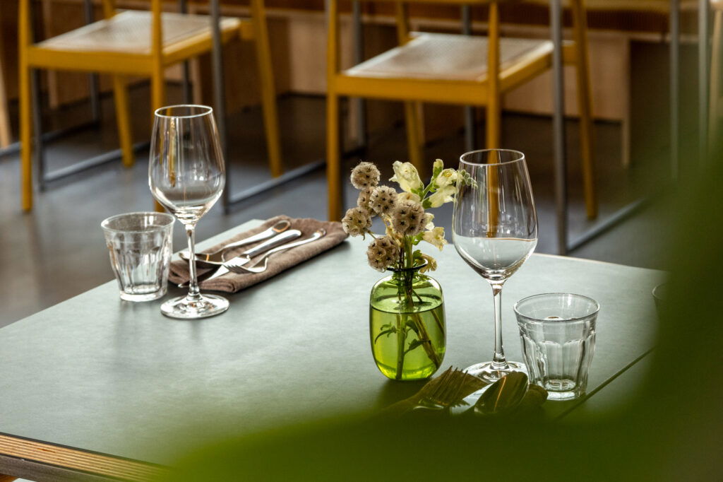 „Elegant gedeckter Restauranttisch mit zwei Weingläsern, Wassergläsern, Besteck auf einer braunen Serviette und einer kleinen Vase mit weißen Blumen; im Hintergrund gelbe Stühle in einem modernen Innenraum.“