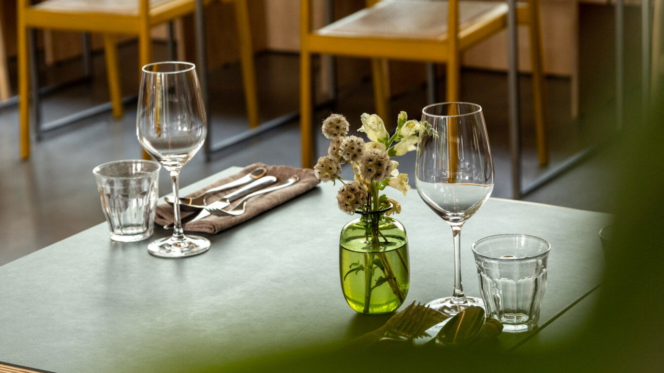 „Elegant gedeckter Restauranttisch mit zwei Weingläsern, Wassergläsern, Besteck auf einer braunen Serviette und einer kleinen Vase mit weißen Blumen; im Hintergrund gelbe Stühle in einem modernen Innenraum.“