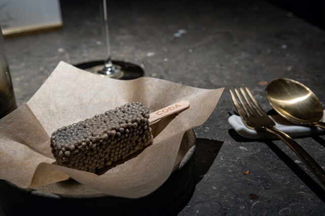 Gourmet-Dessert am Stiel im Fine-Dining-Restaurant in Berlin: ein rechteckiges Eis mit perlartiger Textur auf braunem Papier serviert, daneben elegantes goldenes Besteck