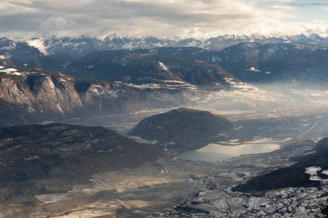Weitwinkelaufnahme des Etschtals mit dem Kalterer See und umliegenden Hügeln im Vordergrund; dahinter breite Bergketten mit Schnee und teils diffusem Licht.