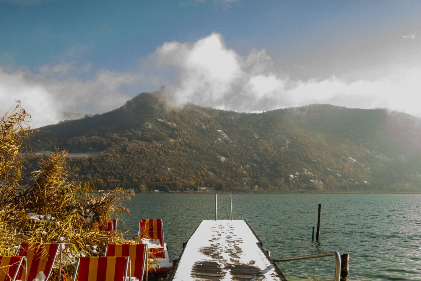Schmaler verschneiter Steg mit Spuren im Schnee, rechts und links bunte Liegestühle, dahinter der Kalterer See und bewaldete Hügel mit etwas Schnee.