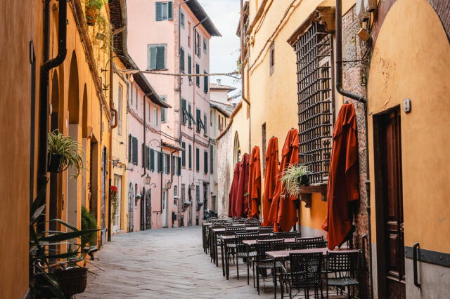 Schmale, gepflasterte Gasse in einer italienischen Altstadt mit warm gelb- und rosafarbenen Hausfassaden; rechts stehen leere Café-Tische mit geschlossenen roten Sonnenschirmen, links lehnt ein Fahrrad an der Wand. Die Gasse führt leicht gebogen auf weitere historische Gebäude zu, überragt von einem Bogen im Vordergrund.