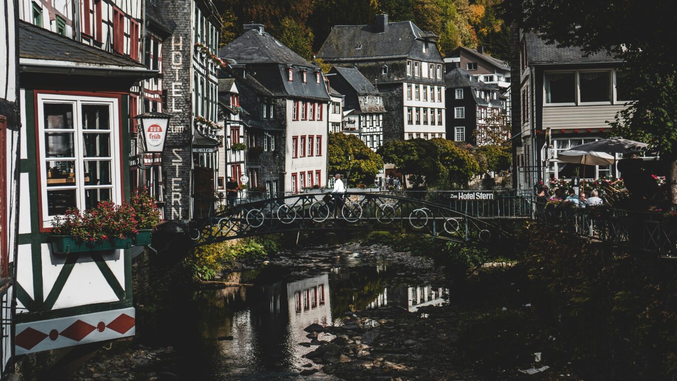 Historische Fachwerkhäuser im Zentrum von Monschau, dicht gedrängt entlang eines kleinen Flusses, mit einer filigranen Fußgängerbrücke im Vordergrund und herbstlich bewaldeten Hügeln im Hintergrund. Ein schönes Ausflugsziel von NRW.