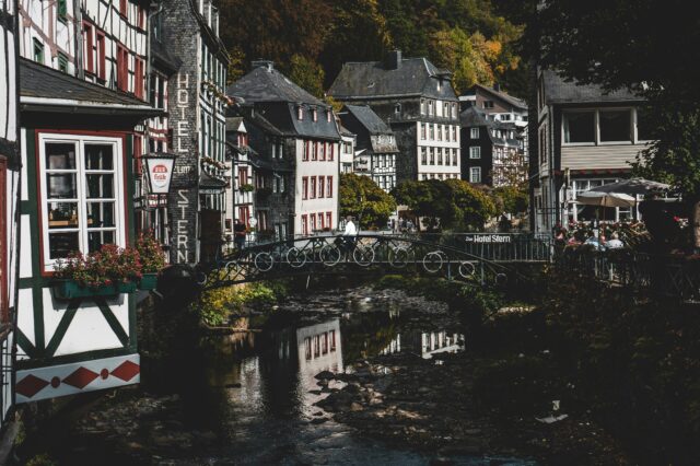Historische Fachwerkhäuser im Zentrum von Monschau, dicht gedrängt entlang eines kleinen Flusses, mit einer filigranen Fußgängerbrücke im Vordergrund und herbstlich bewaldeten Hügeln im Hintergrund. Ein schönes Ausflugsziel von NRW.
