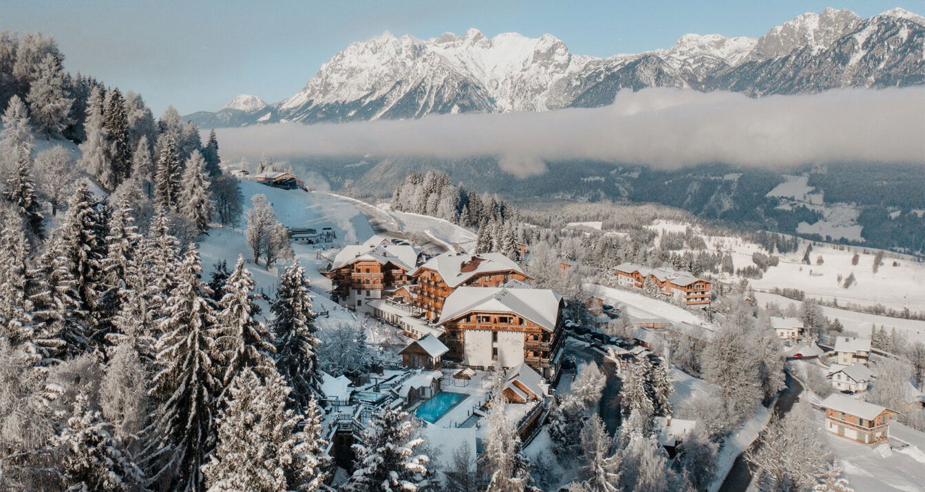 Luftaufnahme eines verschneiten Alpenpanoramas mit dem Natur- und Wellnesshotel Hoflehner in der Bildmitte. Mehrere holzverkleidete Gebäude liegen eingebettet zwischen schneebedeckten Tannen am Hang. Im Vordergrund ist ein beheizter Außenpool zu sehen. Im Hintergrund ragen mächtige, schneebedeckte Berge in den klaren Winterhimmel, während eine tiefliegende Wolkendecke teilweise das Tal verdeckt.