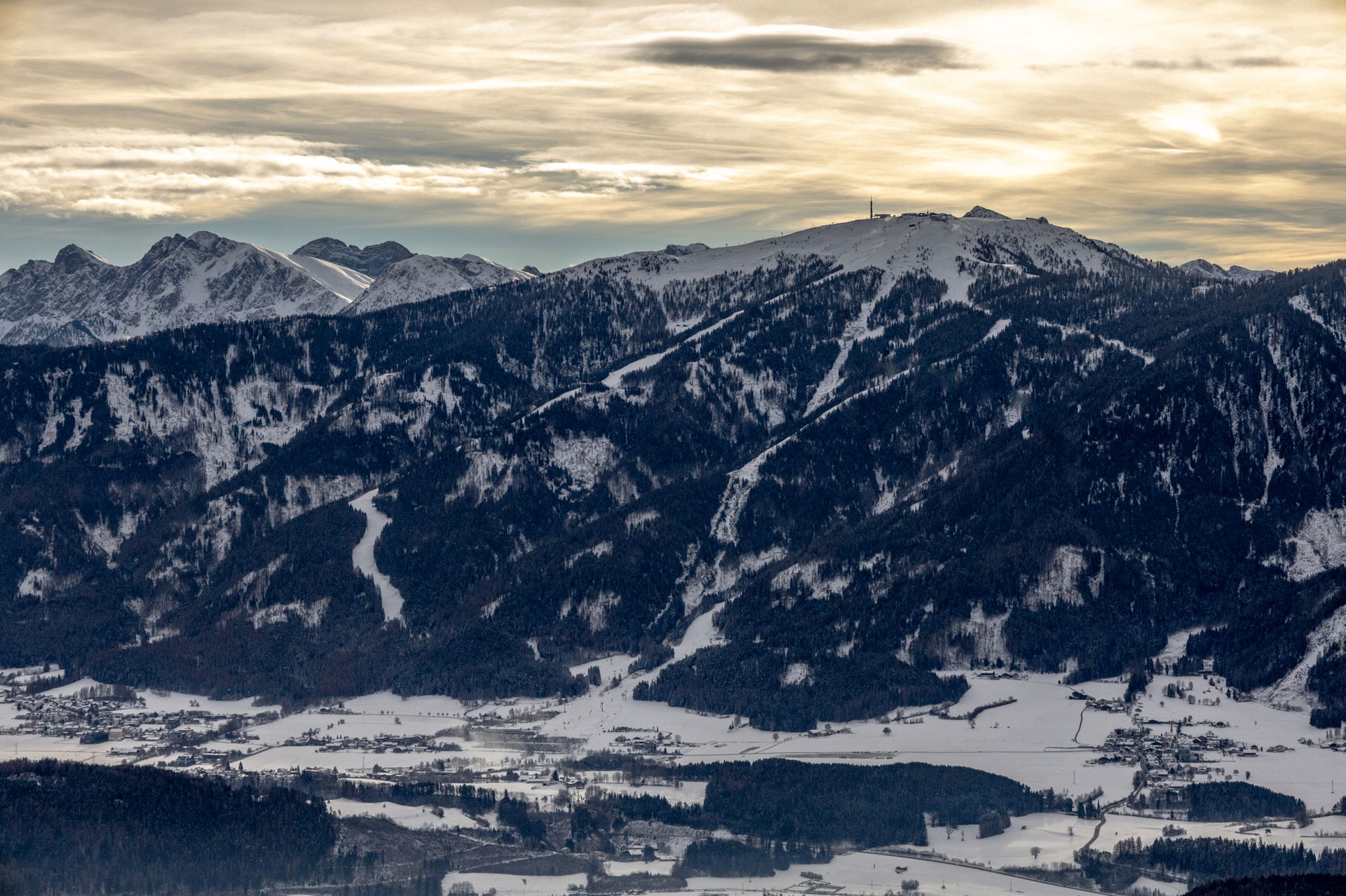 Winterliche Berglandschaft im Pustertal mit schneebedeckten Hängen, dunklen Waldflächen und markanten Gipfeln im Hintergrund. Im Tal liegen verstreute Dörfer und Felder unter einer dünnen Schneedecke. Der Himmel ist von sanftem Abend- oder Morgengold beleuchtet und teils von Wolken überzogen