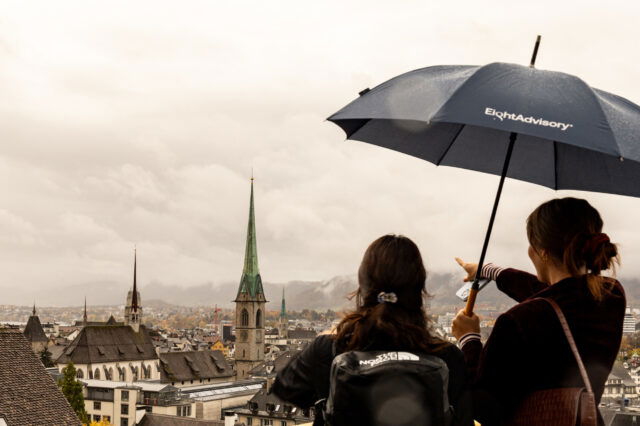 Zwei Personen stehen unter einem Regenschirm auf einem Aussichtspunkt in Zürich und blicken über die Altstadtdächer hinweg auf die markante grüne Kirchturmspitze des Fraumünsters; eine der Personen zeigt in die Ferne, während der Himmel grau und regnerisch ist.