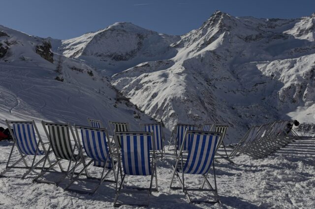 Eine winterliche Berglandschaft mit schneebedeckten Gipfeln unter blauem Himmel. Im Vordergrund stehen mehrere gestreifte Liegestühle im Schnee, daneben zwei Wintersportler*innen mit Ausrüstung. Die Szene wirkt sonnig, ruhig und einladend.