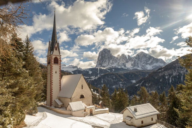 Winterlandschaft in Südtirol mit der verschneiten St.-Jakobs-Kirche auf einer Anhöhe des Grödnertals. Vor dem Panorama ragen die schneebedeckten Gipfel der Geislergruppe in den Himmel. Rund um die Kirche liegen Waldhänge und einzelne Höfe, während Sonnenstrahlen durch die Wolken brechen und die Szene warm beleuchten.