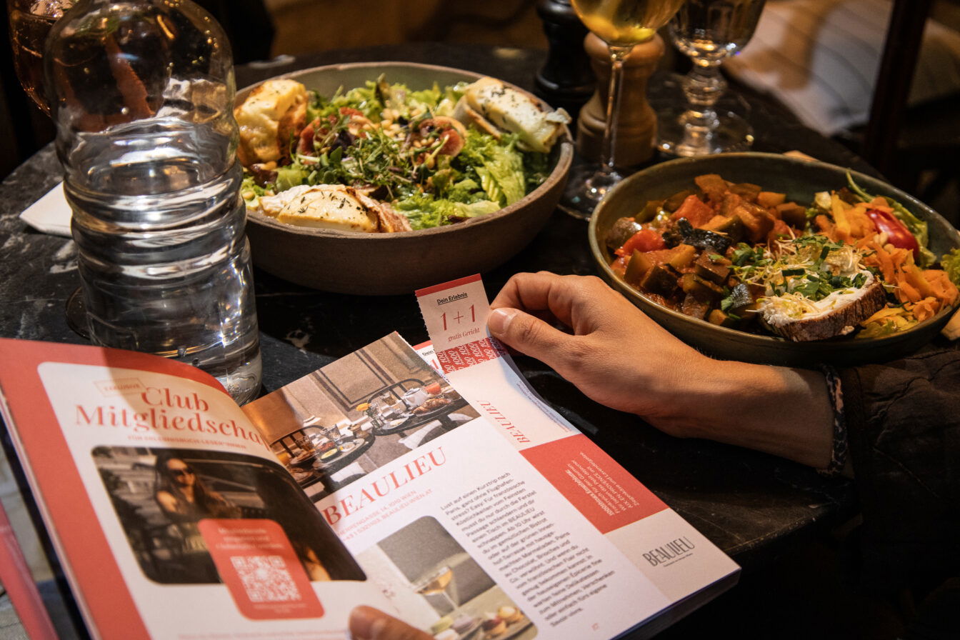 Zwei angerichtete Gerichte auf einem Restauranttisch, daneben ein aufgeschlagenes Genussheft mit Gutschein und eine Hand, die einen 1+1-Voucher hält.