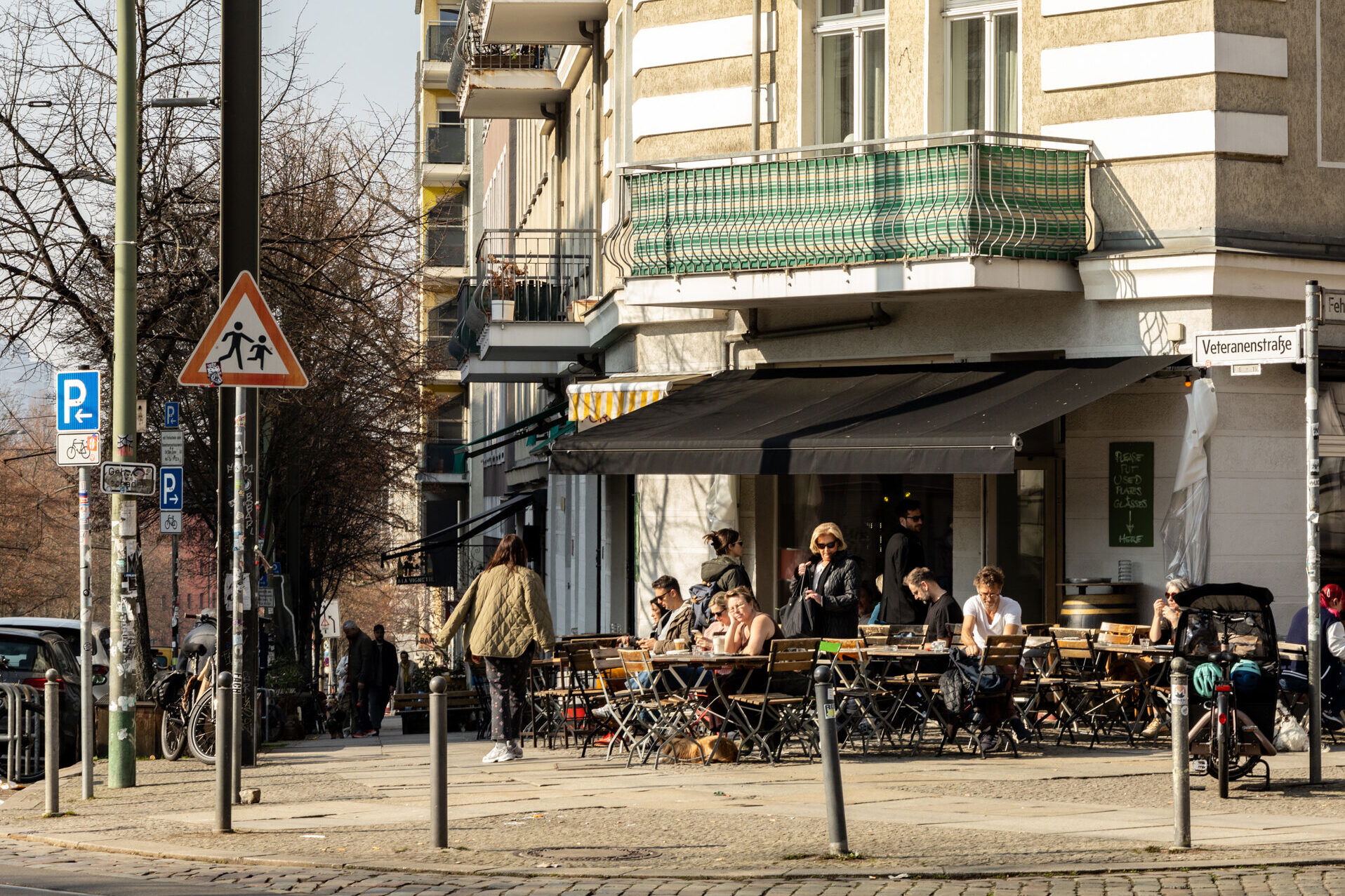 Ein typischer Berliner Straßenzug mit einer einladenden Café- und Bar-Szene. Die Szene spielt sich an der Veteranenstraße ab, wo Menschen das sonnige Wetter genießen und sich an den Tischen eines Straßencafés niederlassen. Schwarze Markisen spenden Schatten, während die Gäste in entspannter Atmosphäre sitzen, plaudern und ihr Essen oder Getränke genießen.
