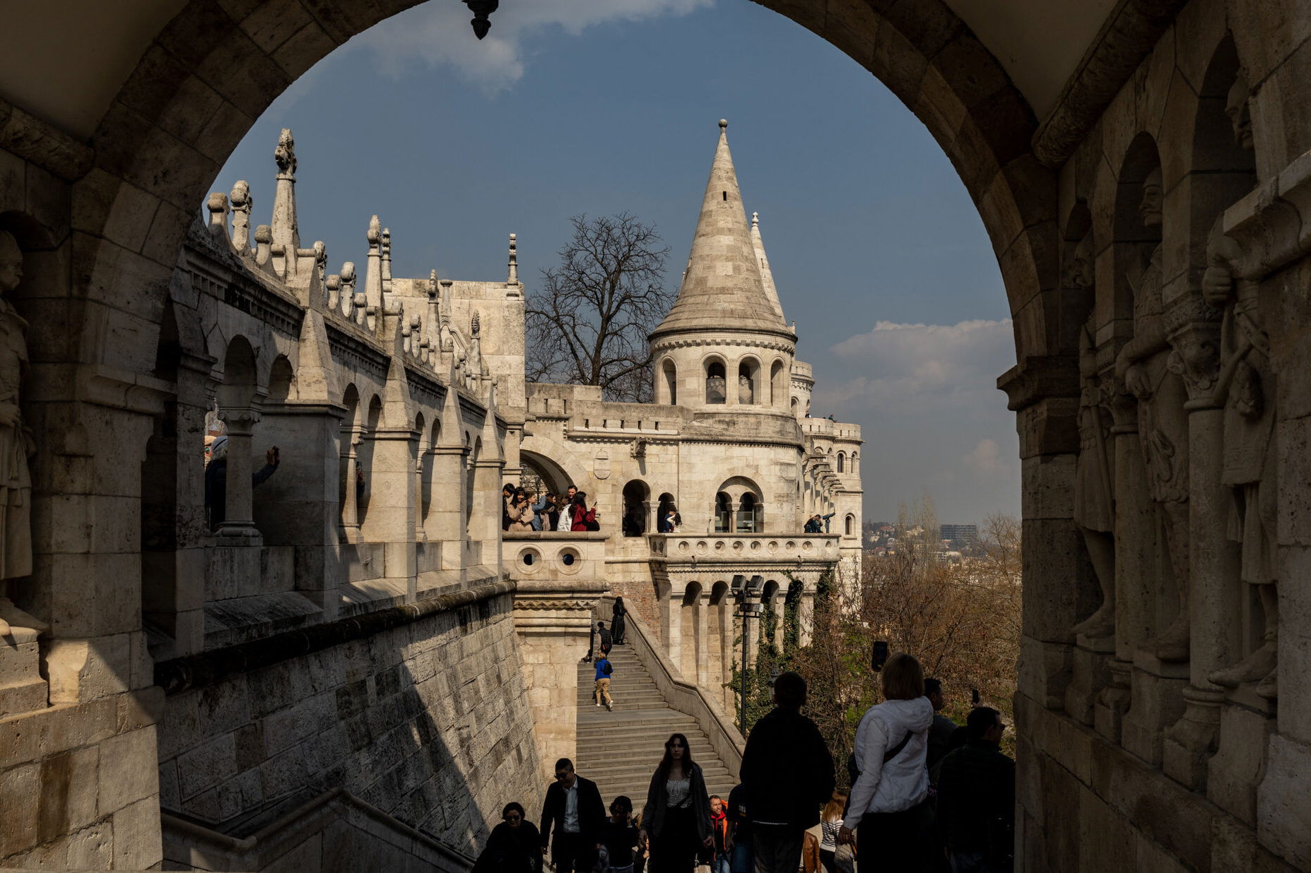 Tourist*innen steigen die Treppen zur Fischerbastei in Budapest hinauf. Der Blick führt durch einen steinernen Torbogen mit verzierten Figurenreliefs auf die romantische Architektur der Bastei mit ihren Türmchen, Rundbögen und Balustraden. Das Sonnenlicht hebt die Details des hellen Sandsteins hervor. Die Fischerbastei zählt zu den beliebtesten Sehenswürdigkeiten Budapests mit einzigartigem Ausblick über die Stadt und die Donau.
