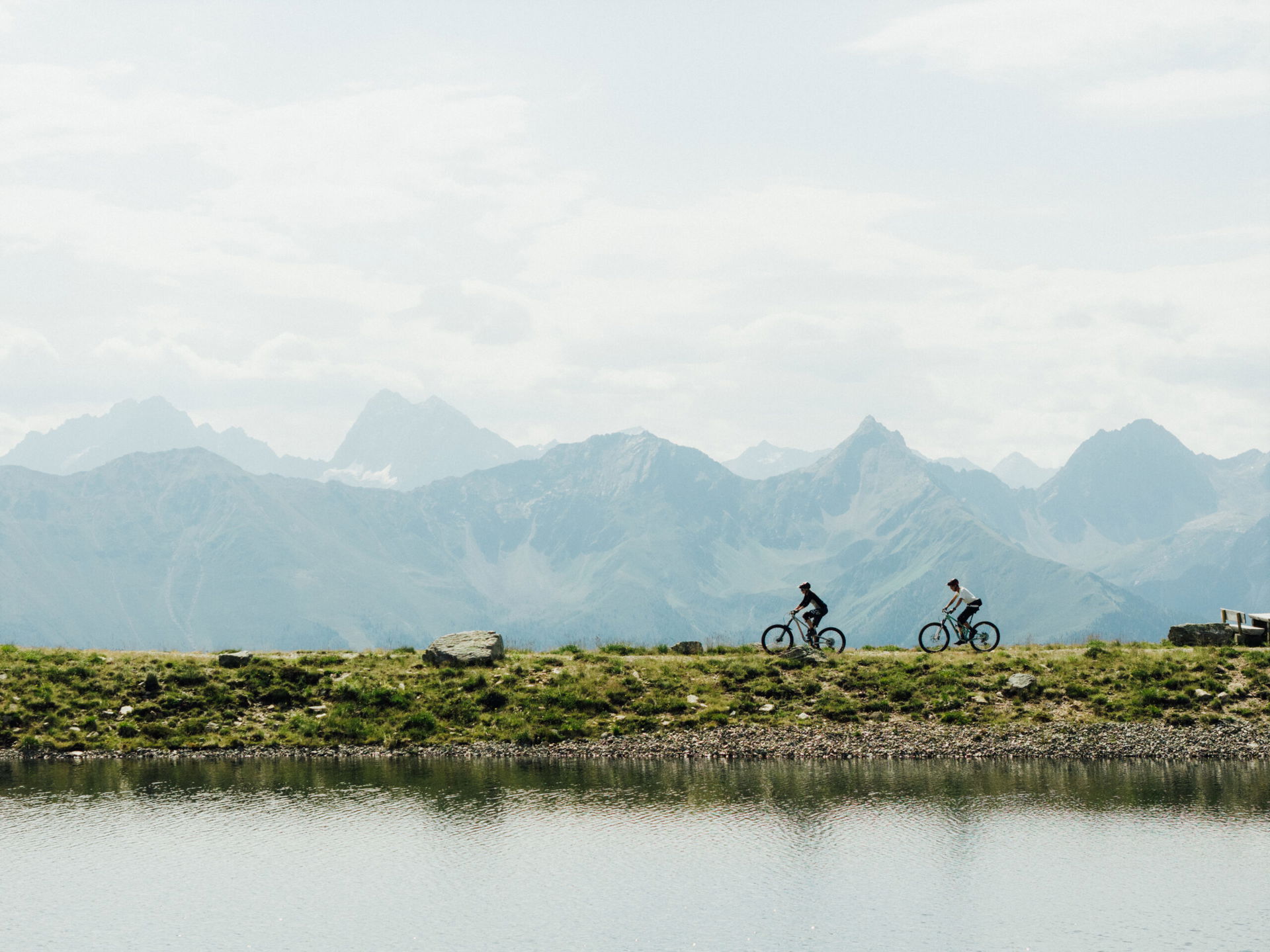 Zwei Radfahrer fahren an einem See entlang, dahinter ist ein vernebeltes Bergpanorama zu sehen.