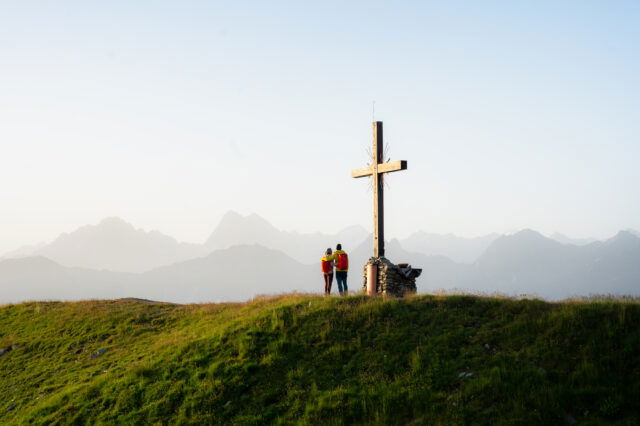 Zwei Personen stehen auf einem Berg beim Gipfelkreuz und bewundern die Berglandschaft.