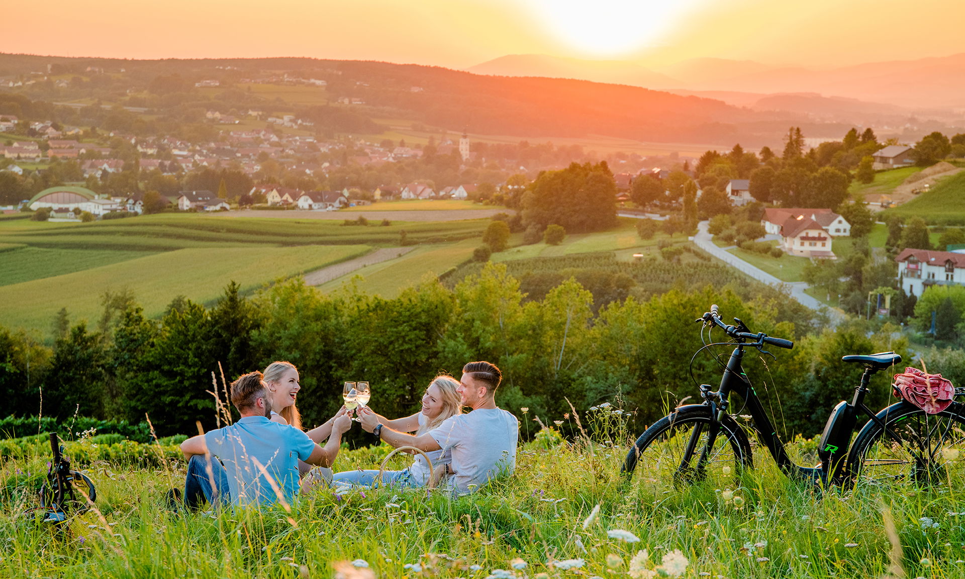 Hügelige Landschaft mit Weingarten bei Sonnenuntergang. Im Vordergrund stoßt eine Gruppe von Menschen mit Wiengläsern an.