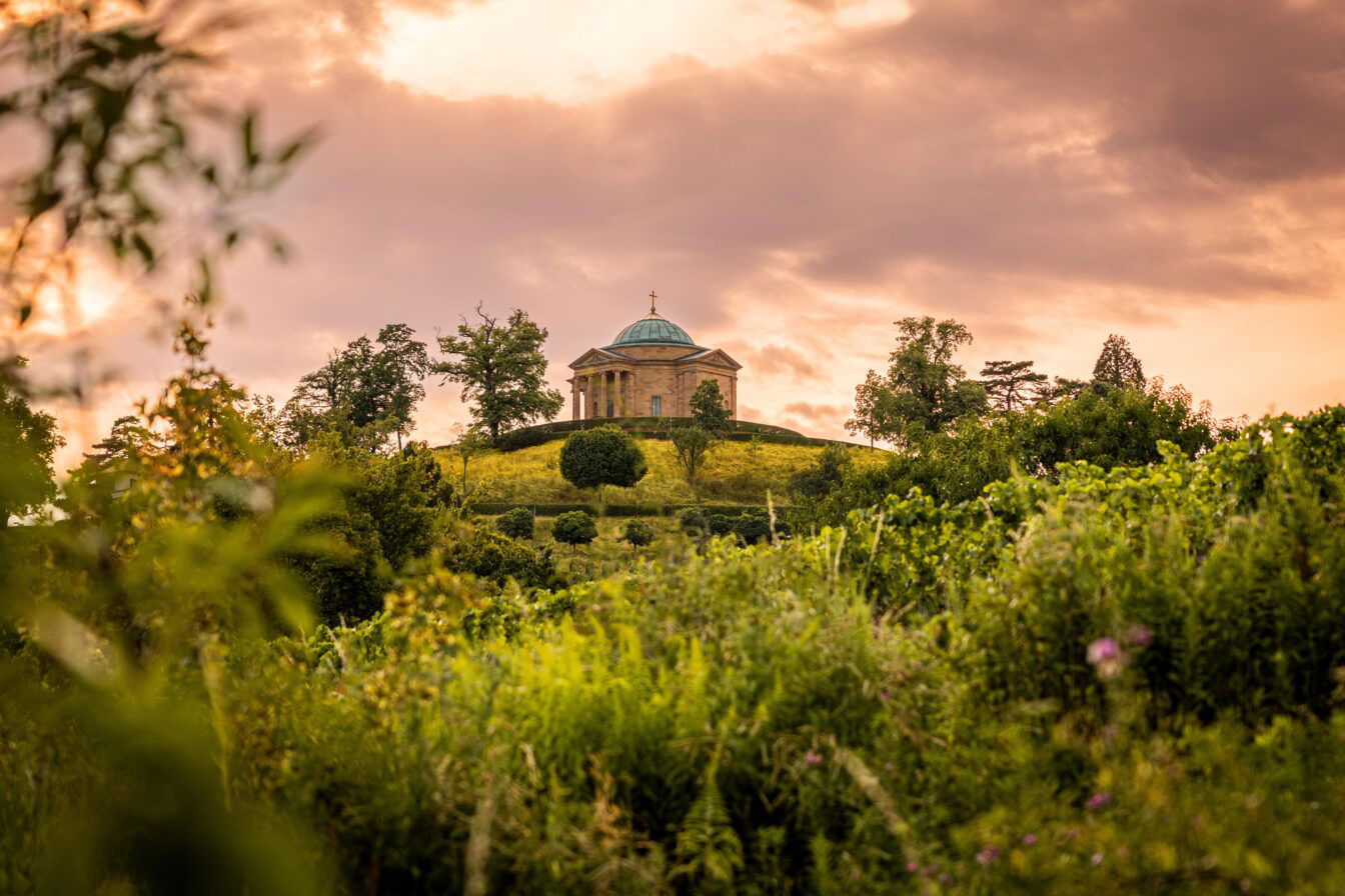 Kleine Kapelle in grüner Umgebung, imposanter Himmel. Es ist die Grabkapelle auf dem Württemberg.
