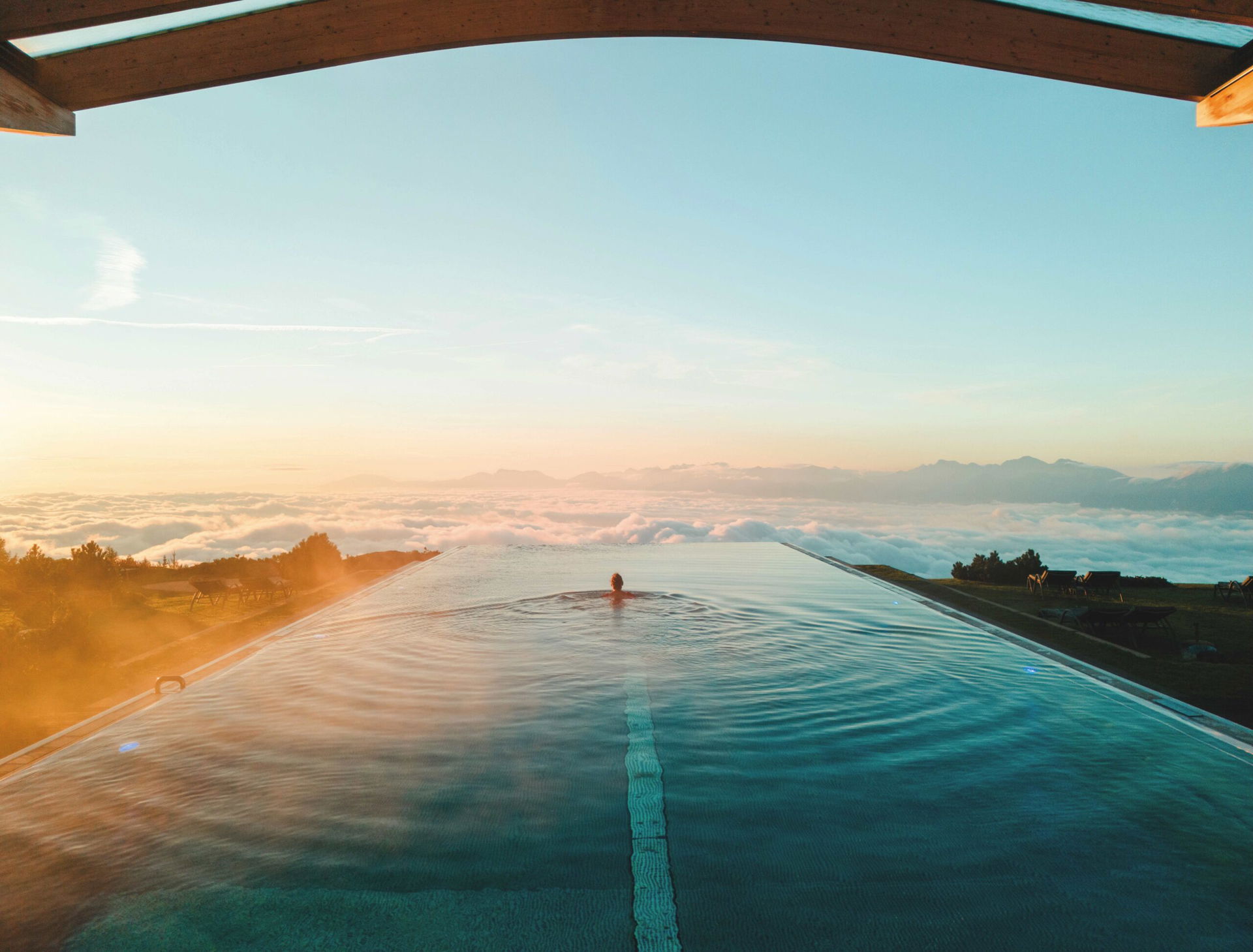 Person schwimmt im Unendlichpool am Mountain Resort Feuerberg auf den Rand zu, dahinter sieht man nur Wolken und Bergspitzen.