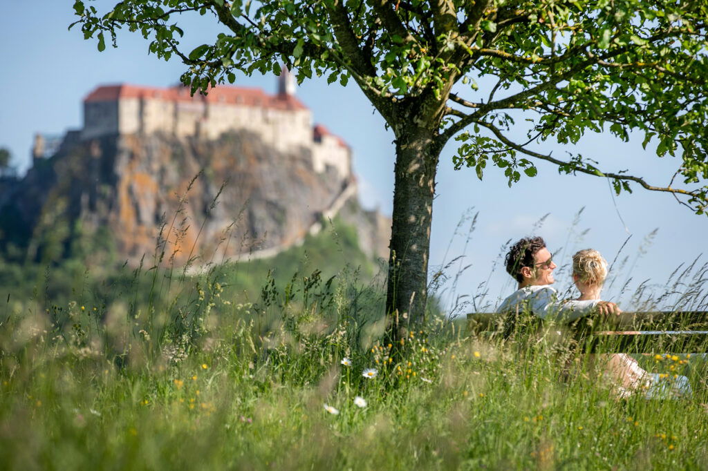 Blick auf eine Burg mit einem Pärchen im Vordergrund, das auf einer Bank unter einem Baum sitzt.