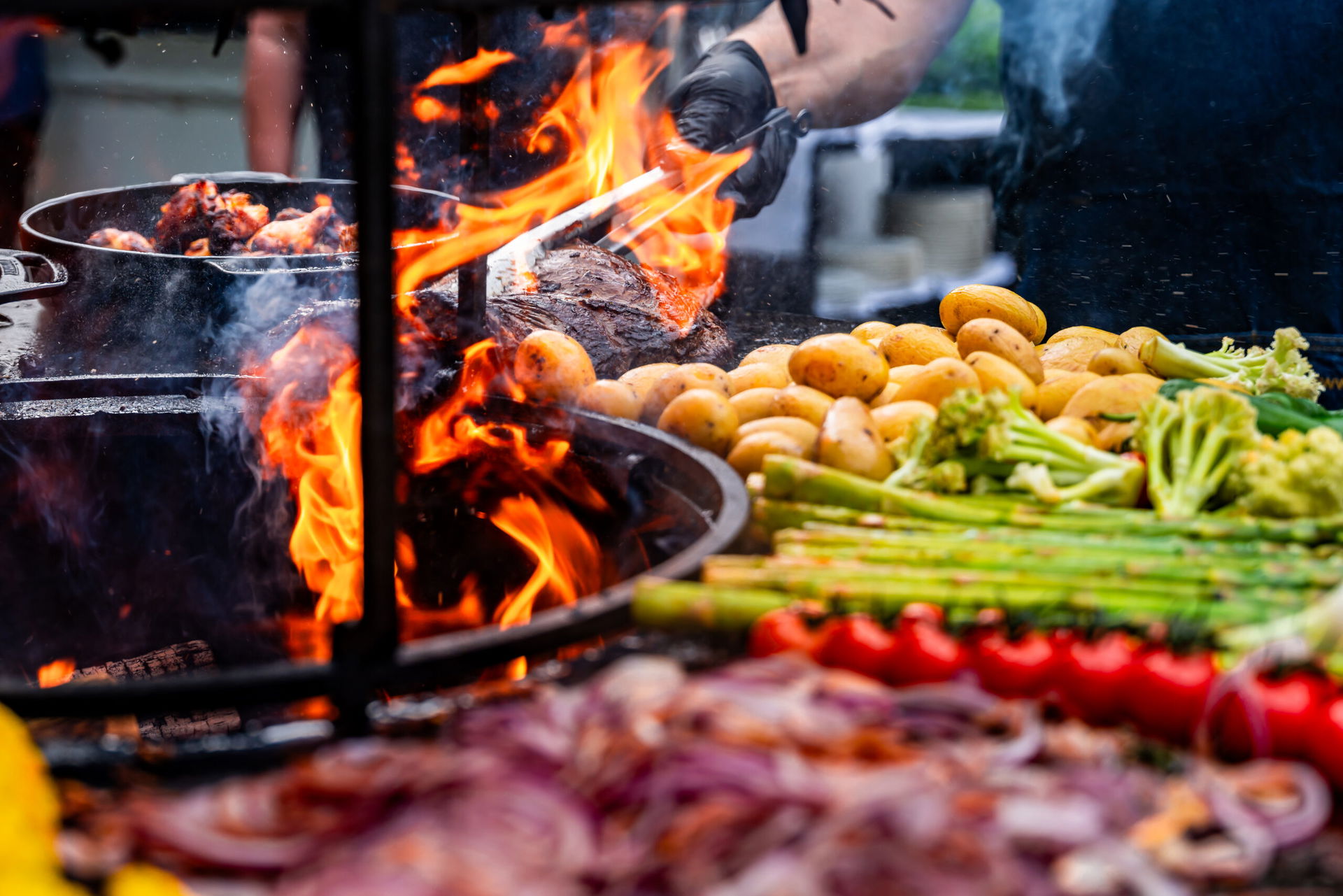 Man sieht einen Grill mit Feuer und vielen Zutaten: Erdäpfel, Spargel, Fleisch, Zwiebel, Tomaten. 
Es ist das Küste BBQ beim Strandhotel Zingst.