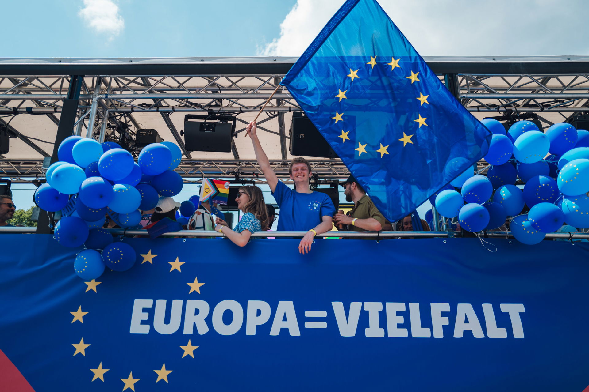 Drei Personen sind auf einer blauen Bühne zu sehen, ein junger Mann schwenkt eine Europäische Flagge, unter ihm ist der Schriftzug "Europa=Vielfalt" aufgedruckt und neben ihm sind viele blaue Luftballons mit den Europäischen Sternen zu sehen.