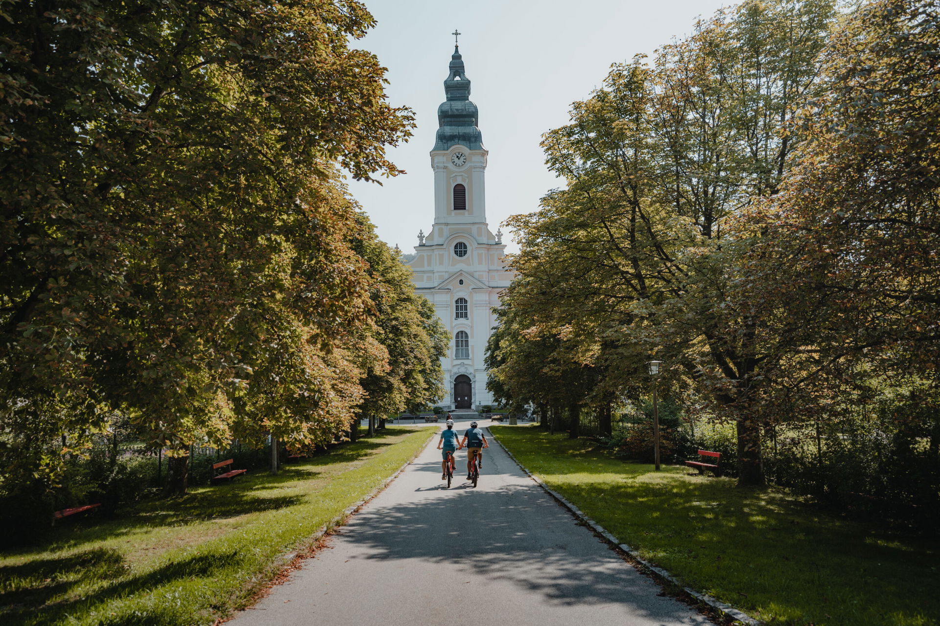 Zwei Personen fahren die Sauwald Panorama-Runde und steuern auf Stift Engelszell, eine große, weiße Kirche zu. © CM Visuals 