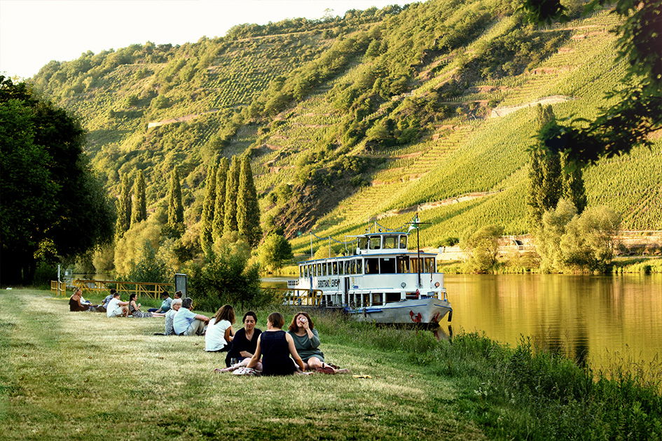 Ein Schiff fährt im Sommer über den Mosel Fluss in Rheinland Pfalz. Am Ufer sitzen Menschen und trinken Wein.